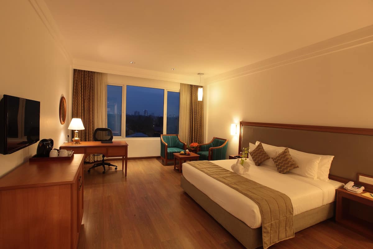 Habitación de hotel con una cama grande, un escritorio con lámpara, dos sillas, televisión y vista a la ciudad al anochecer.