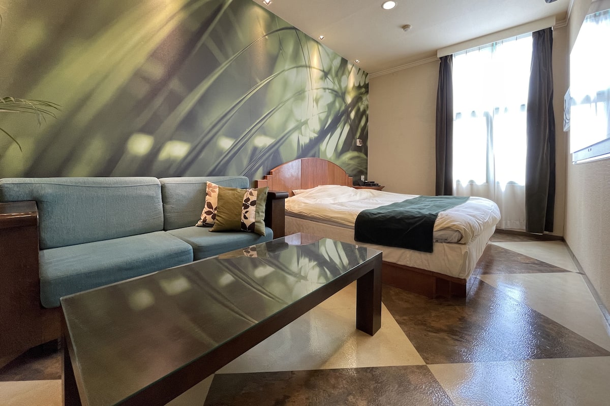 A modern hotel room with a sofa, a bed, a glass coffee table, and a large window with curtains.