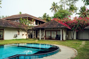 A swimming pool with a curved edge, surrounded by a grassy lawn and a building with a tiled roof. - Cranganor History Cafe Riverside Chateau (North Paravur)