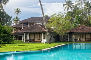 A poolside view of a resort with a large pool, two-story buildings with tiled roofs, and lush greenery including palm trees. - Cranganor History Cafe Riverside Chateau (North Paravur)