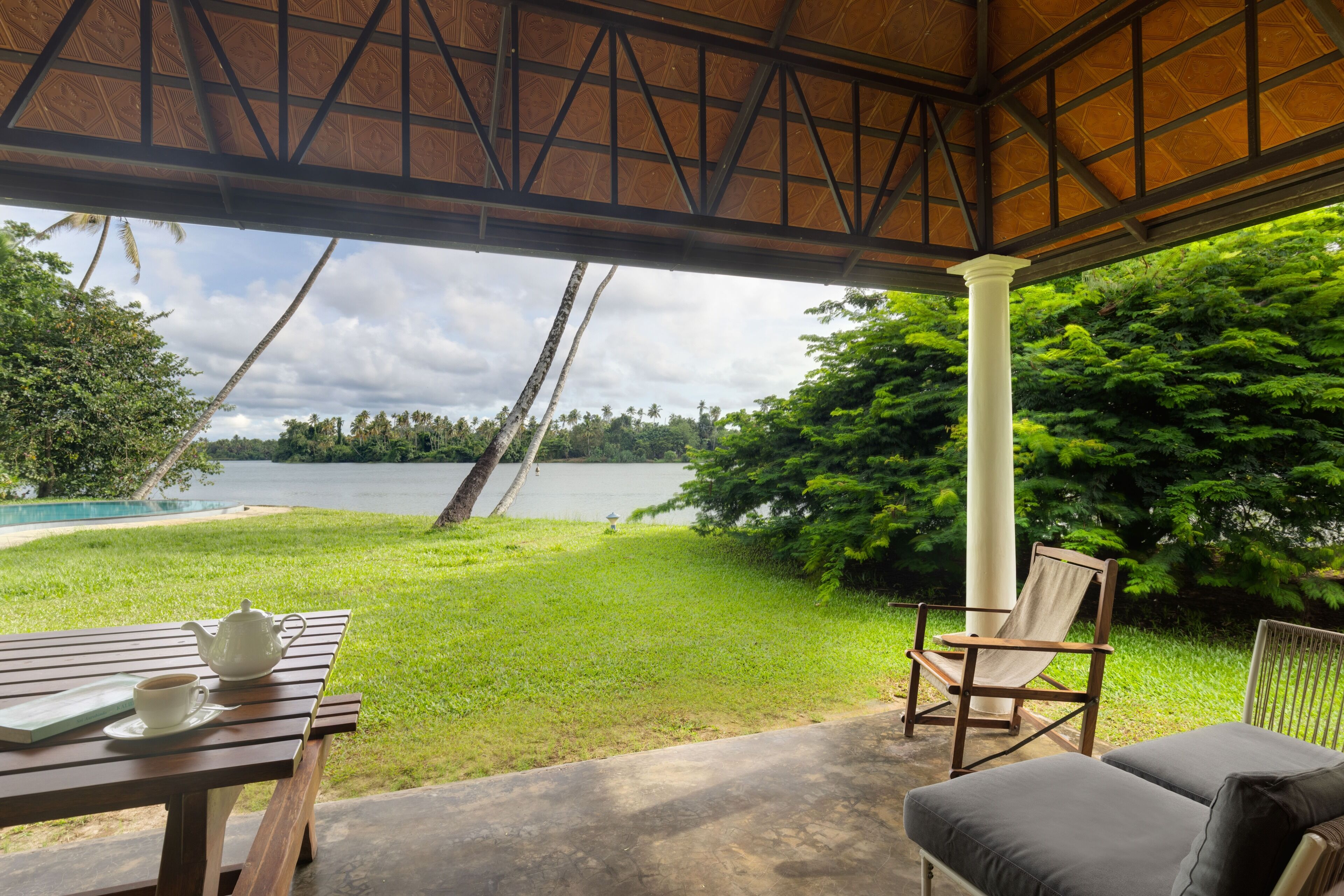 A covered outdoor seating area with a table and chairs, overlooking a lake and lush greenery.