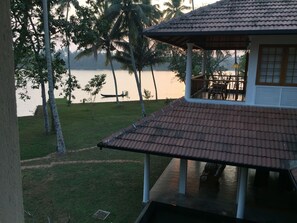 A building with a tiled roof, a balcony with chairs, and a view of a calm body of water with palm trees in the background. - Cranganor History Cafe Riverside Chateau (North Paravur)