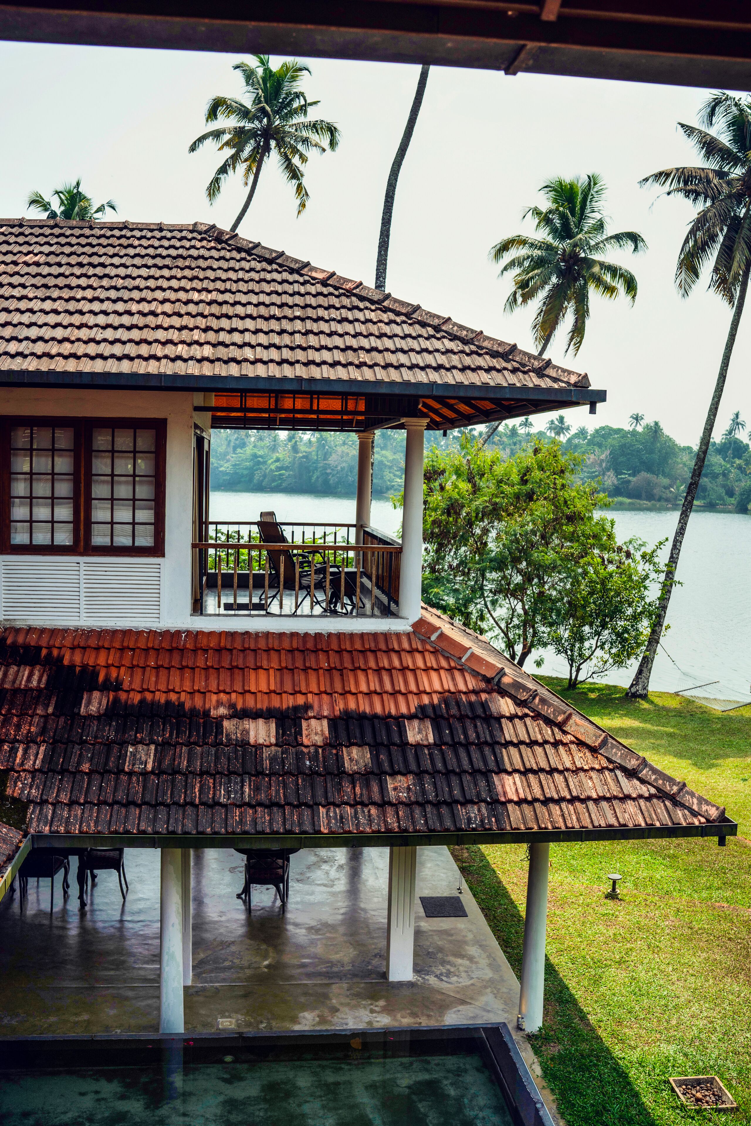 A house with a tiled roof, a balcony with chairs, and a view of a lake with palm trees.