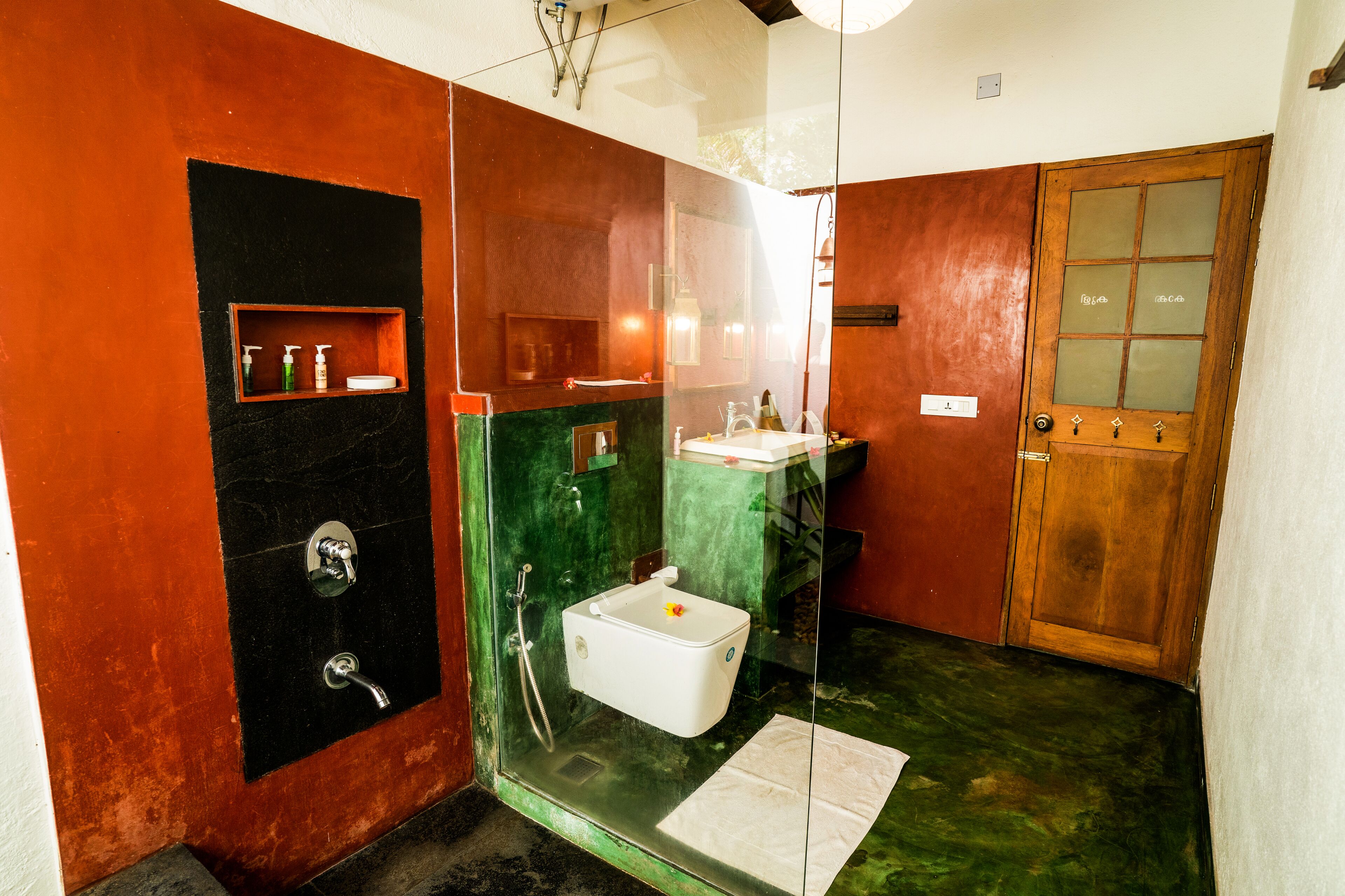 A compact bathroom with a glass enclosure, a wooden door, and a small shelf with toiletries.