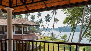 A view from a balcony overlooking a lake with palm trees, a wooden building with a tiled roof, and a grassy area. - Cranganor History Cafe Riverside Chateau (North Paravur)