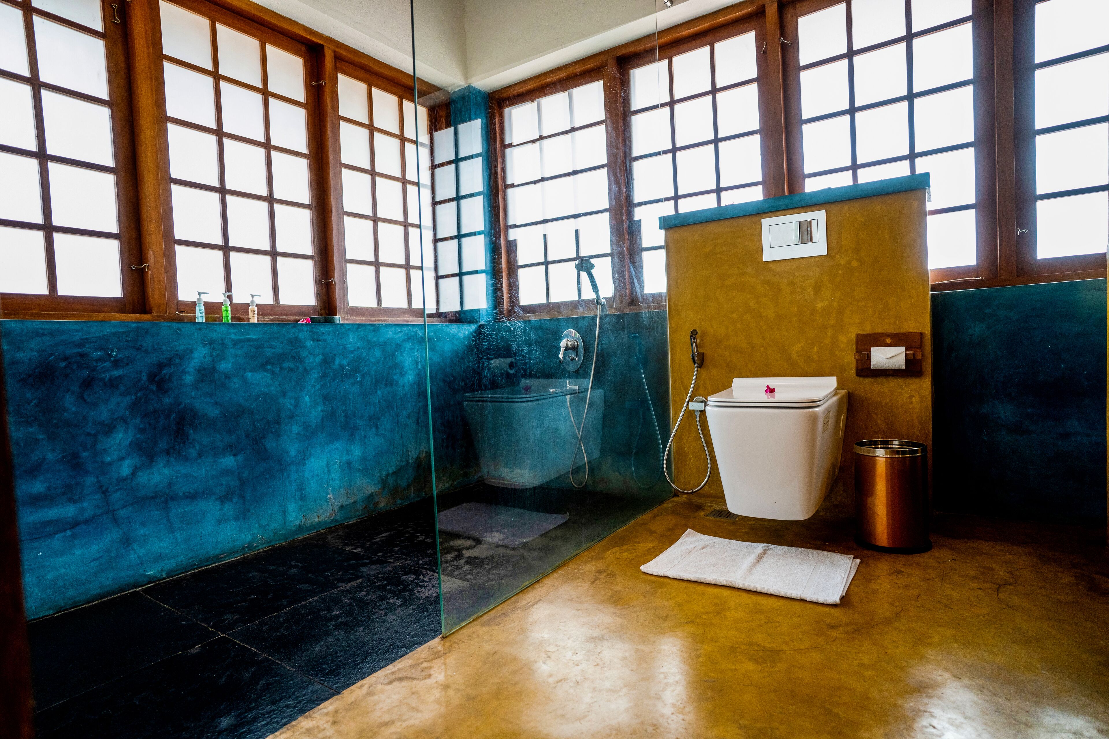 A bathroom with a blue and brown tiled floor, a white toilet, a shower, and a window with wooden frames.