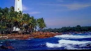 Un faro con torre blanca y parte superior negra, rodeado de palmeras y costa rocosa.