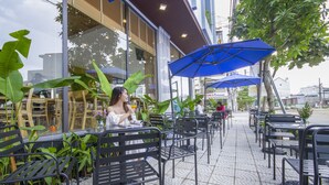 An outdoor cafe with metal chairs and tables, some under blue umbrellas, and large green plants around. - King House Villa Hotel (Da Nang)