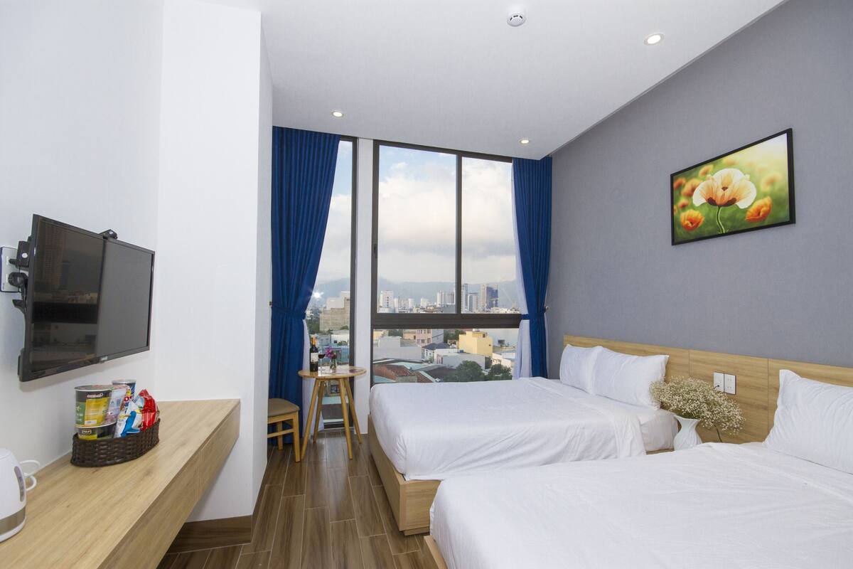 Habitación de hotel con dos camas, televisión, una mesita con aperitivos y vistas a la ciudad a través de amplias ventanas.