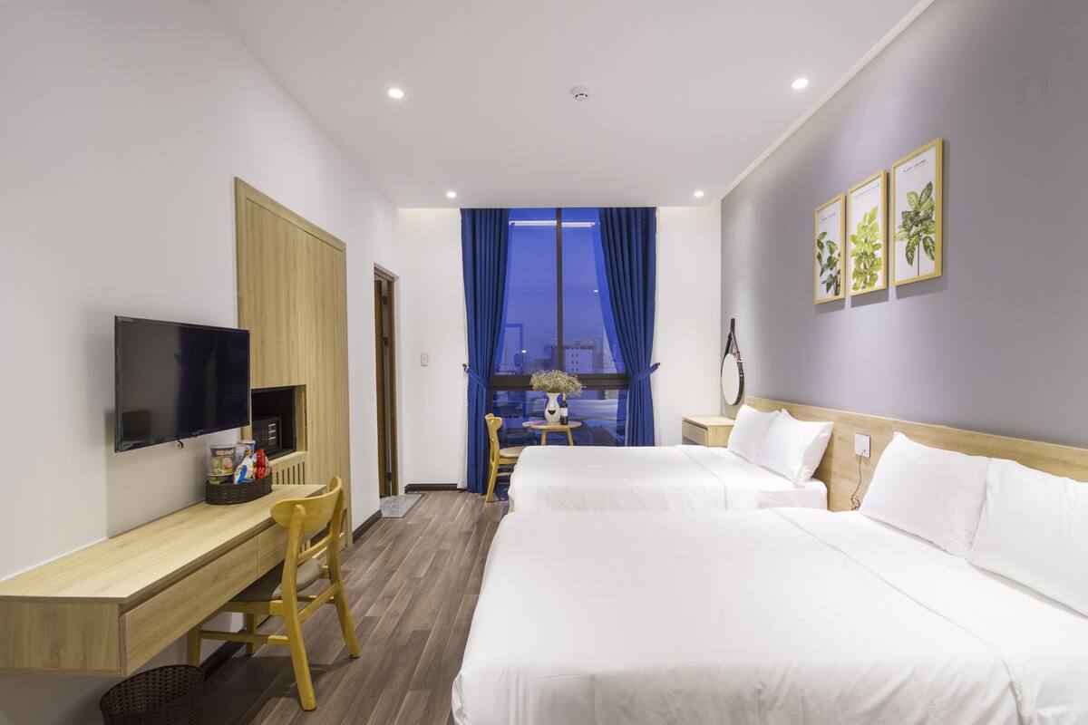 Habitación de hotel con dos camas, un escritorio con televisor, una silla, una mesita y una ventana con cortinas.
