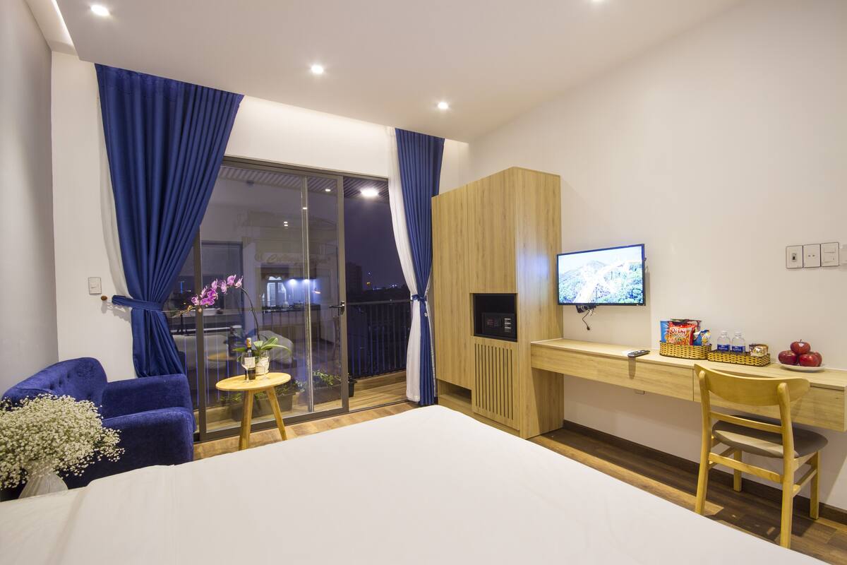 Habitación de hotel moderna con cama, escritorio con computadora, silla, una pequeña mesa con planta y balcón con cortinas.