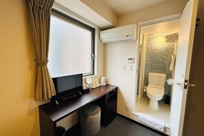 Habitación de hotel compacta con escritorio, televisor, taburete pequeño, ventana con cortinas, unidad de aire acondicionado, inodoro y zona de ducha.