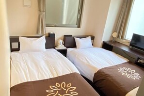 Habitación de hotel con dos camas, cada una con ropa blanca y un cojín decorativo marrón. Hay una ventana con cortinas, una mesita de noche con una lámpara y un televisor sobre un mueble.