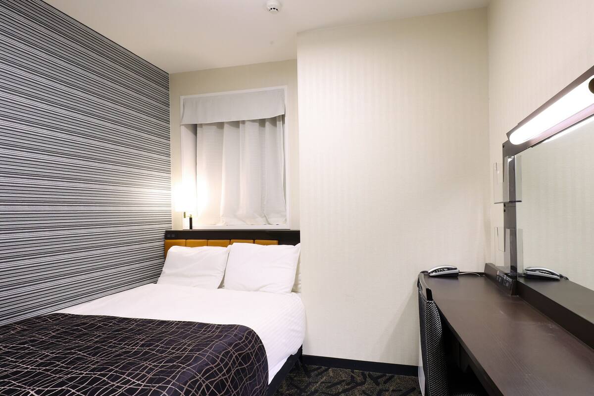 A hotel room with a bed, a desk, a mirror, and a window with curtains.