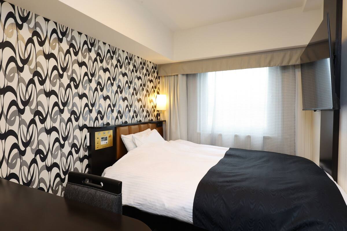 A hotel room with a patterned wallpaper, a bed with white and black bedding, a bedside table, a lamp, and a television mounted on the wall.