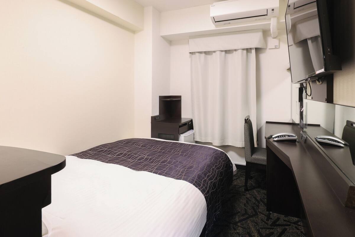 A hotel room with a bed, a desk, a chair, a TV, and an air conditioning unit.