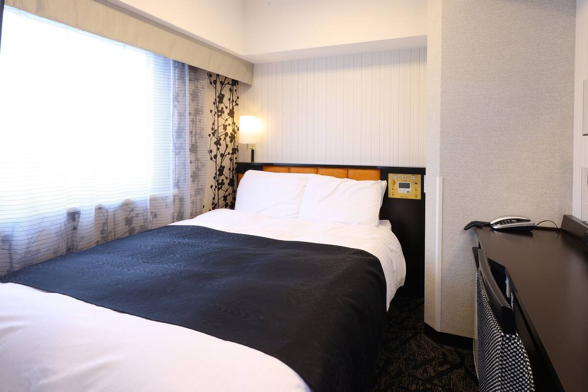 A hotel room with a double bed, a bedside table with a phone, and a window with curtains.