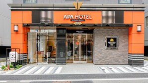 Exterior del APA HOTEL con un diseño moderno, destacando una fachada naranja prominente, una entrada de vidrio amplia y un letrero que indica la dirección.