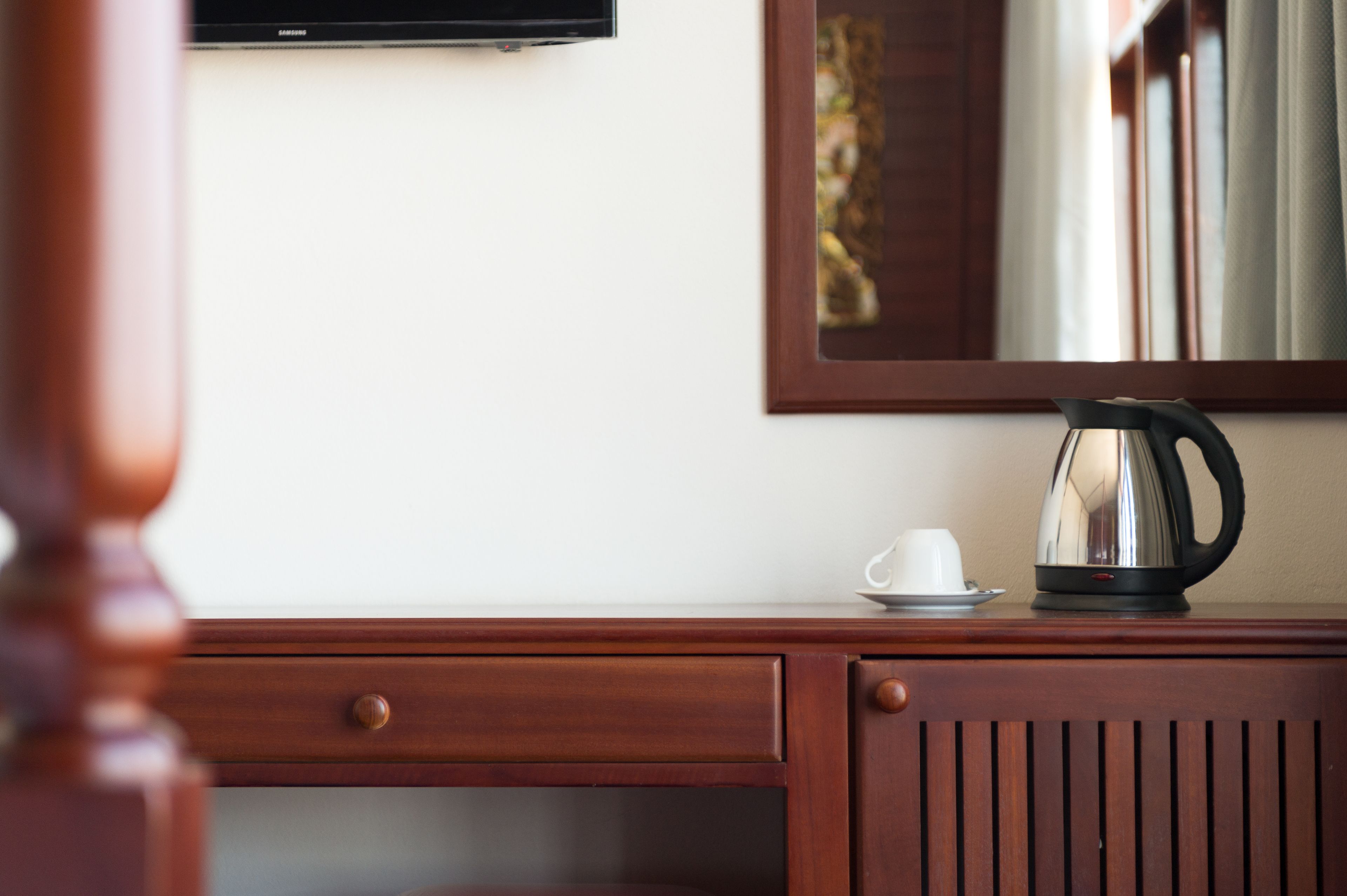 Een houten bureau met een kopje en een waterkoker, een televisie aan de wand bevestigd, en een lijst met een afbeelding erboven.