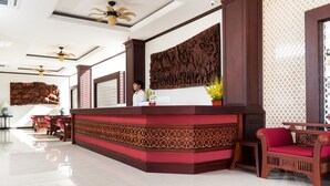 A reception area with a wooden reception desk, a large carved wall, and a seating area with a red cushioned chair. - Manorom Chateau Hotel (Vientiane)