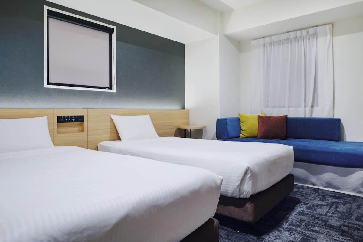 Habitación de hotel con dos camas, una mesita, un sofá y un ventanal con cortinas.