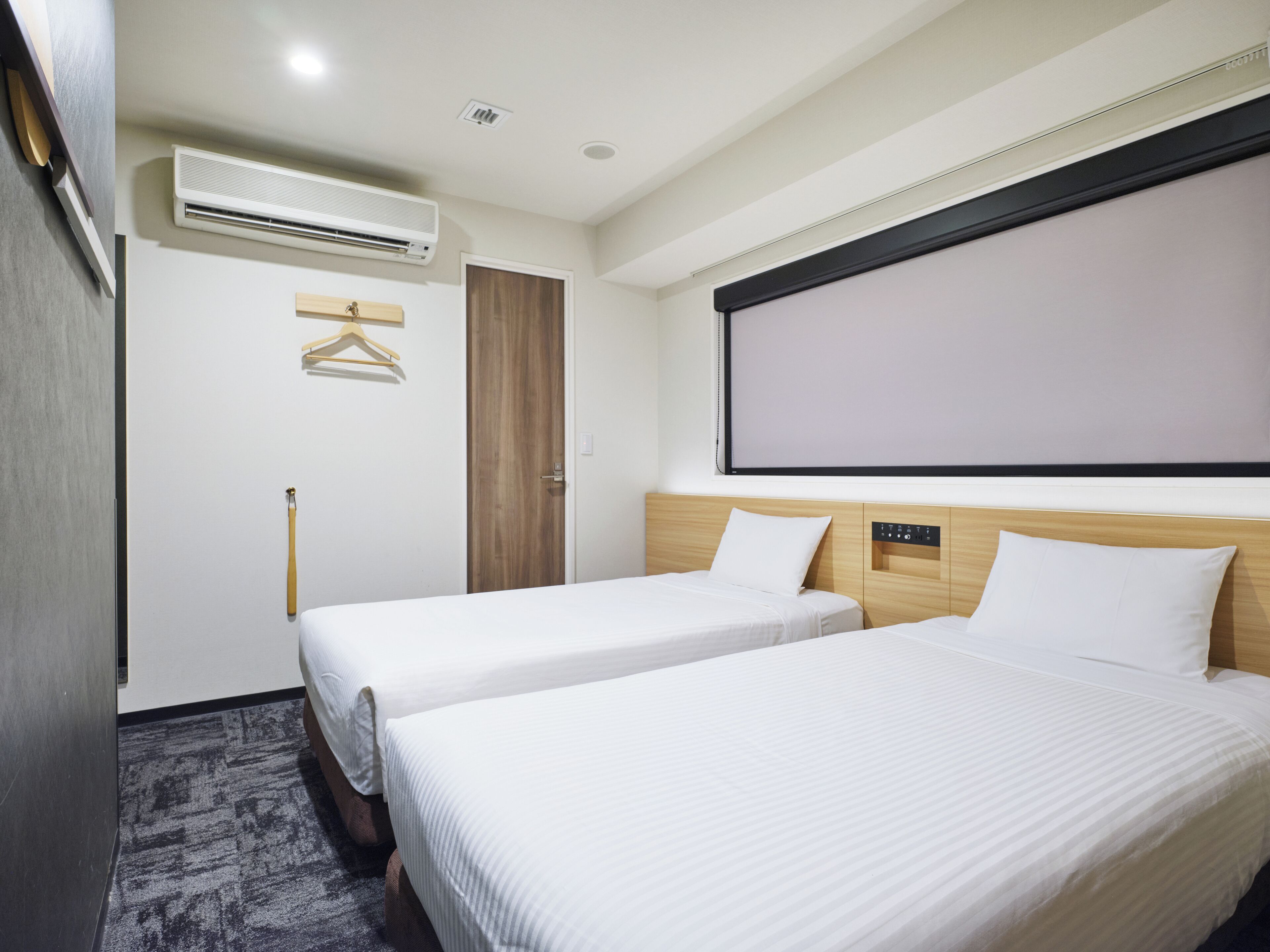 Une chambre d’hôtel avec deux lits, un appareil de climatisation, une porte en bois, un porte-manteau et une grande fenêtre.