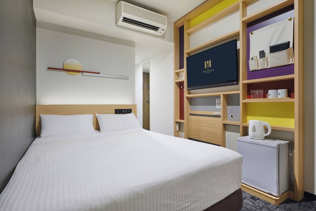 Una habitación de hotel con cama, aire acondicionado, televisión, mini refrigerador y una repisa con tazas y tetera.