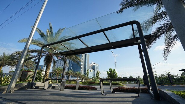 An outdoor seating area with metal benches and a glass canopy, surrounded by palm trees and modern buildings. - The Lively Hotel Kualanamu Airport Medan (Medan)