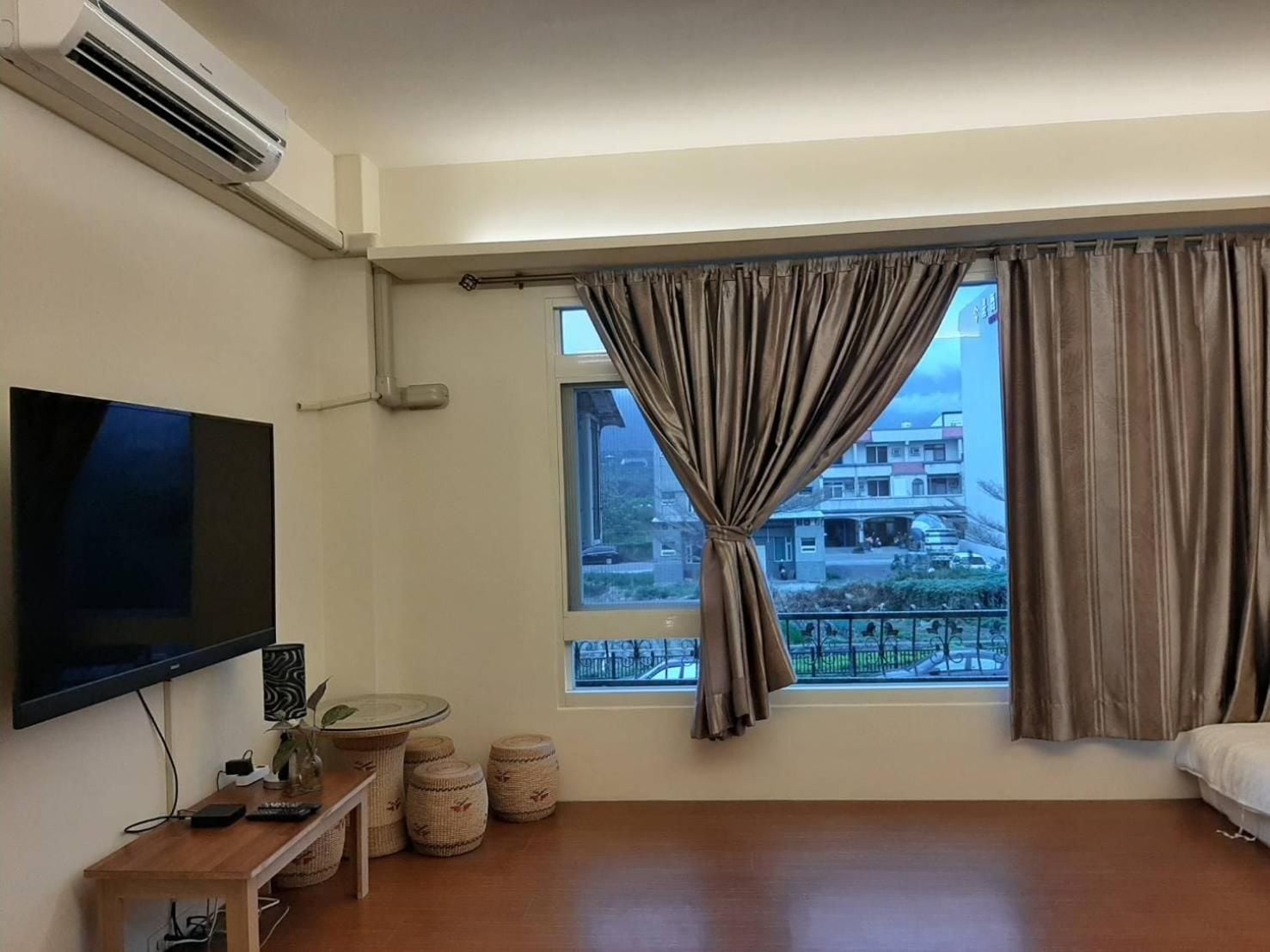 A hotel room with a flat-screen TV, a window with curtains, a wooden floor, and a view of buildings outside.