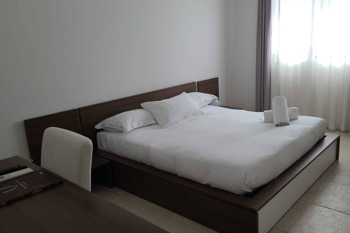 Ein ordentlich bezogenes Bett mit weißen Leinen und Kissen, ein Nachttisch mit einem Laptop und ein Fenster mit Vorhängen.