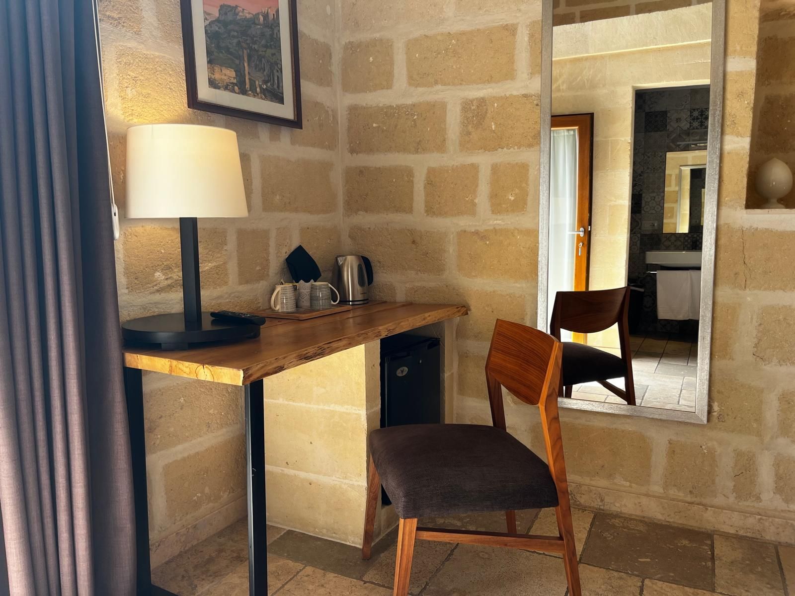 Una habitación con escritorio, silla, lámpara y un espejo que refleja un baño.