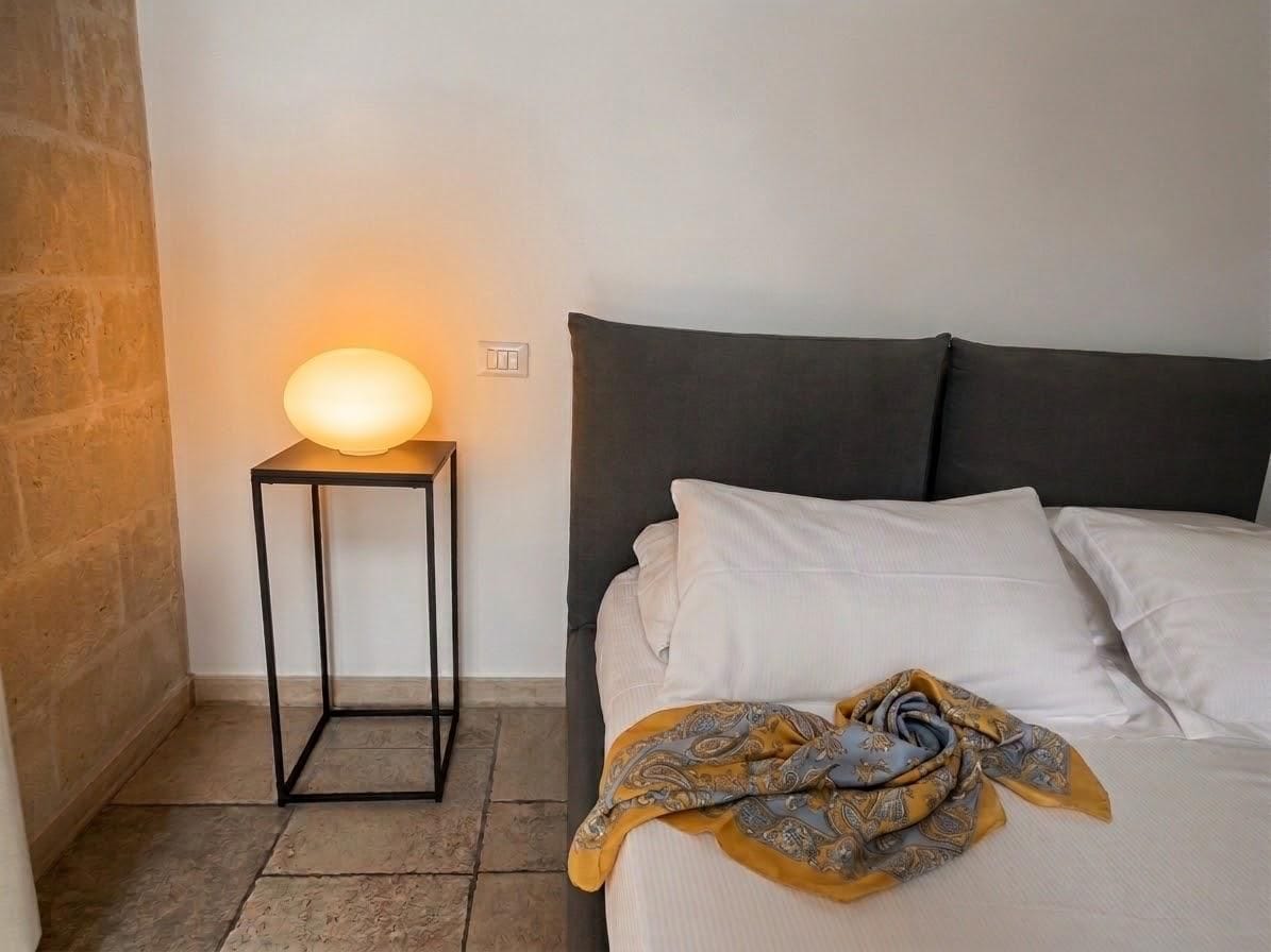 A bed with white linens and a patterned throw blanket, a bedside table with a lamp, and a stone wall.