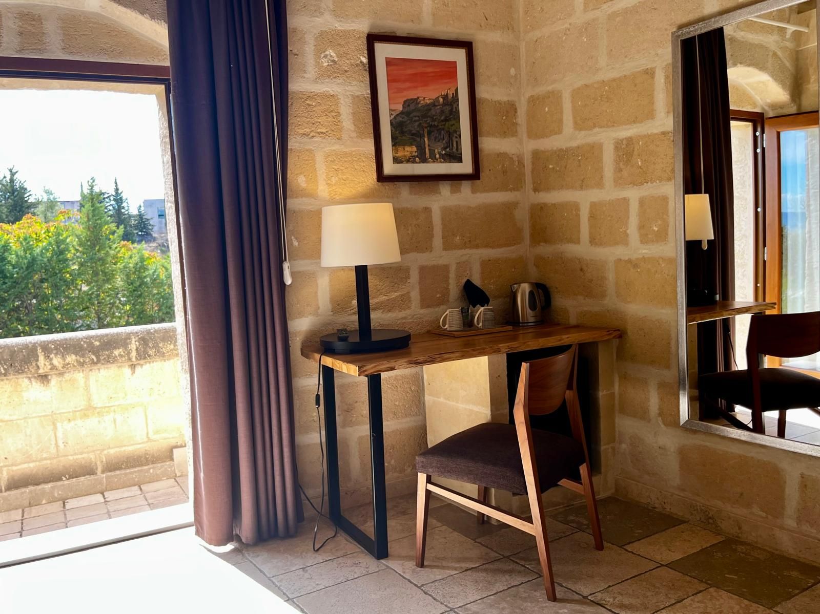 Una habitación con pared de piedra, un escritorio con lámpara, una silla y una ventana con cortinas.
