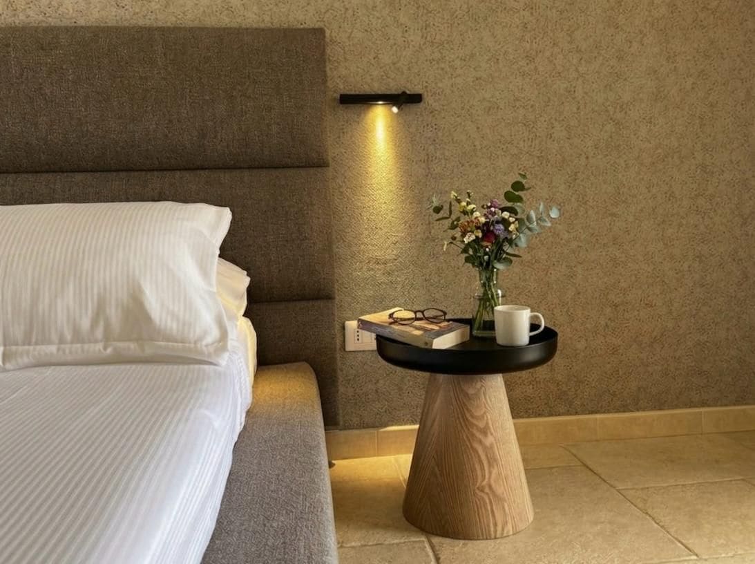 A neatly made bed with white linens, a bedside table with a vase of flowers, a book, and a cup.
