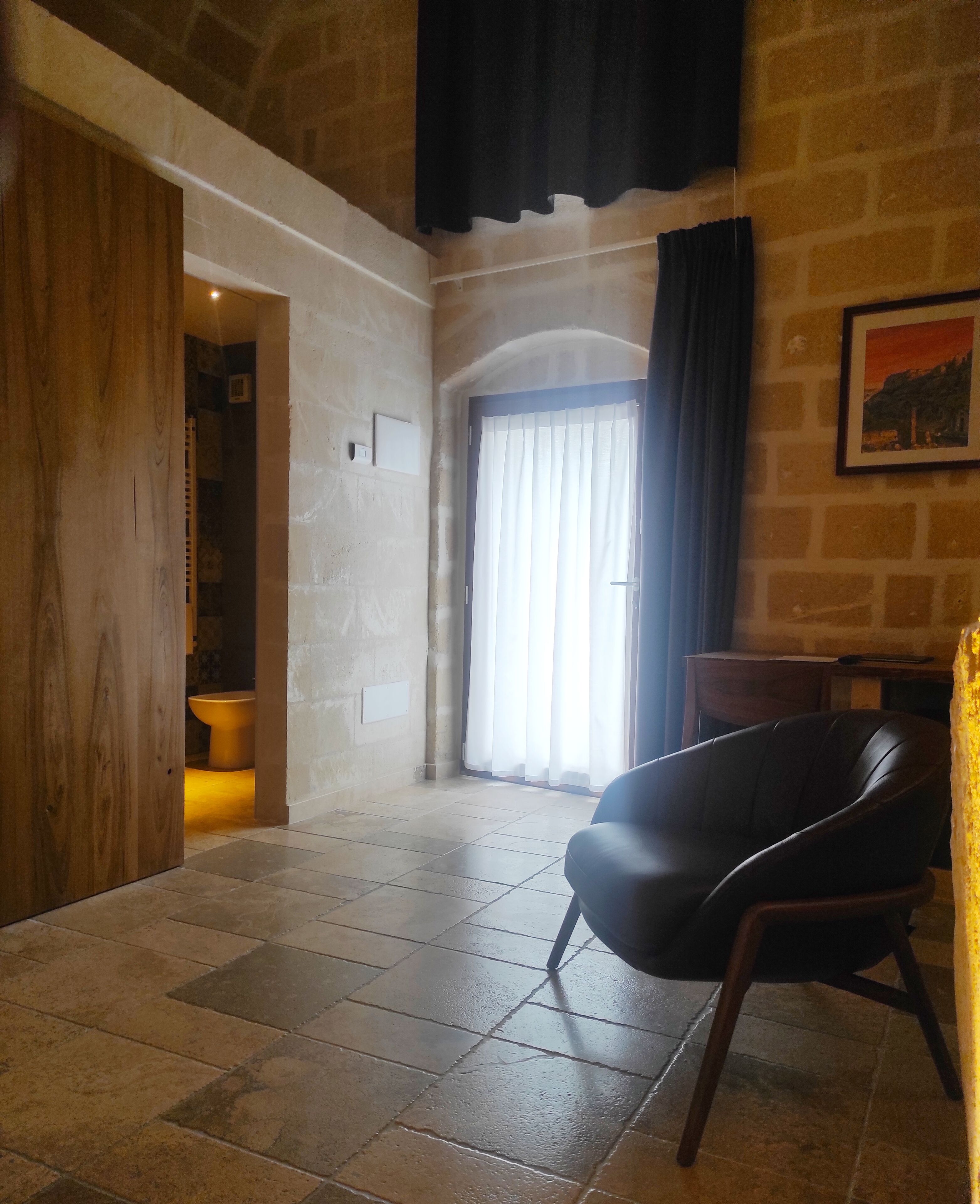 Una habitación amplia con un gran ventanal, una puerta de madera, un sillón de cuero negro y una pared de piedra.