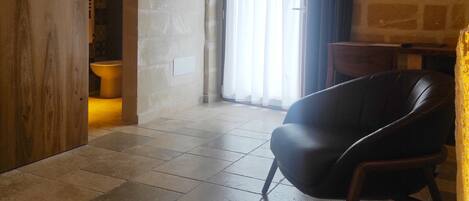 Una habitación amplia con un gran ventanal, una puerta de madera, un sillón de cuero negro y una pared de piedra.