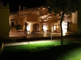 Un'area salotto esterna con tavoli e sedie, un albero e un edificio con archi e facciata in pietra.