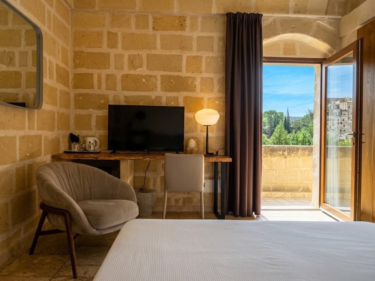 Ein Hotelzimmer mit Backsteinwand, einem Bett, einem Schreibtisch mit Fernseher, einem Stuhl und Blick ins Freie.