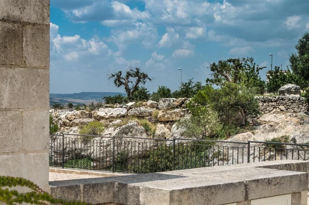 A stone wall with a metal railing in the foreground, a rocky landscape with sparse vegetation in the background, and a clear sky above.