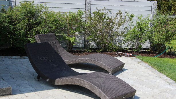 Two outdoor lounge chairs on a paved patio with a hedge and a house in the background. - Gästehaus Zehmerhof (Walpertskirchen)