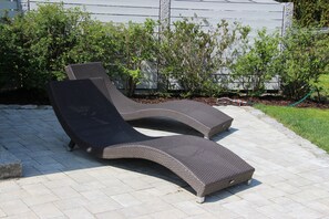 Two outdoor lounge chairs on a paved patio with a hedge and a house in the background. - Gästehaus Zehmerhof (Walpertskirchen)