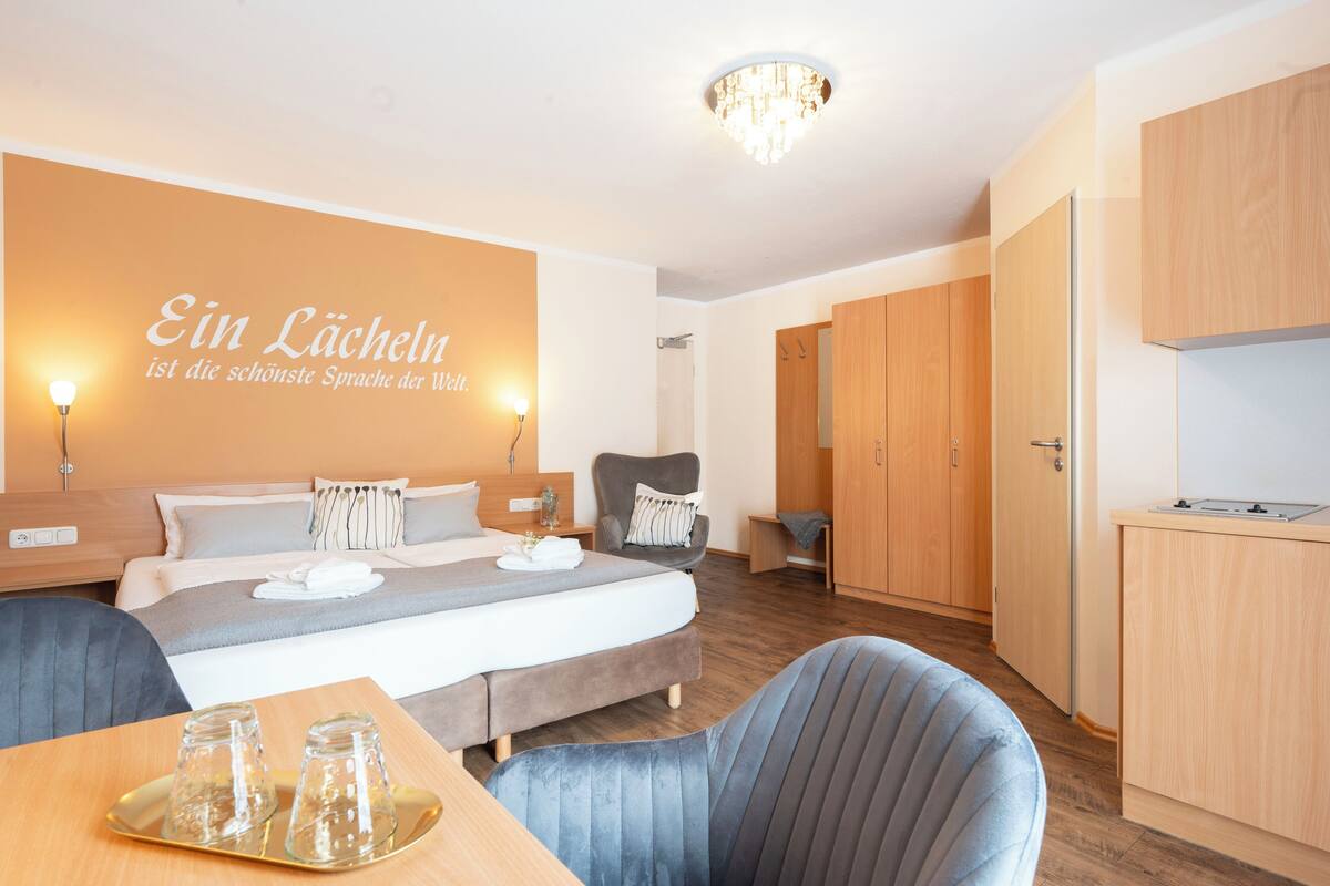 Une chambre d’hôtel moderne, équipée d’un lit, d’une chaise, d’un bureau avec un plateau de verres et d’un mur orné d’une citation.