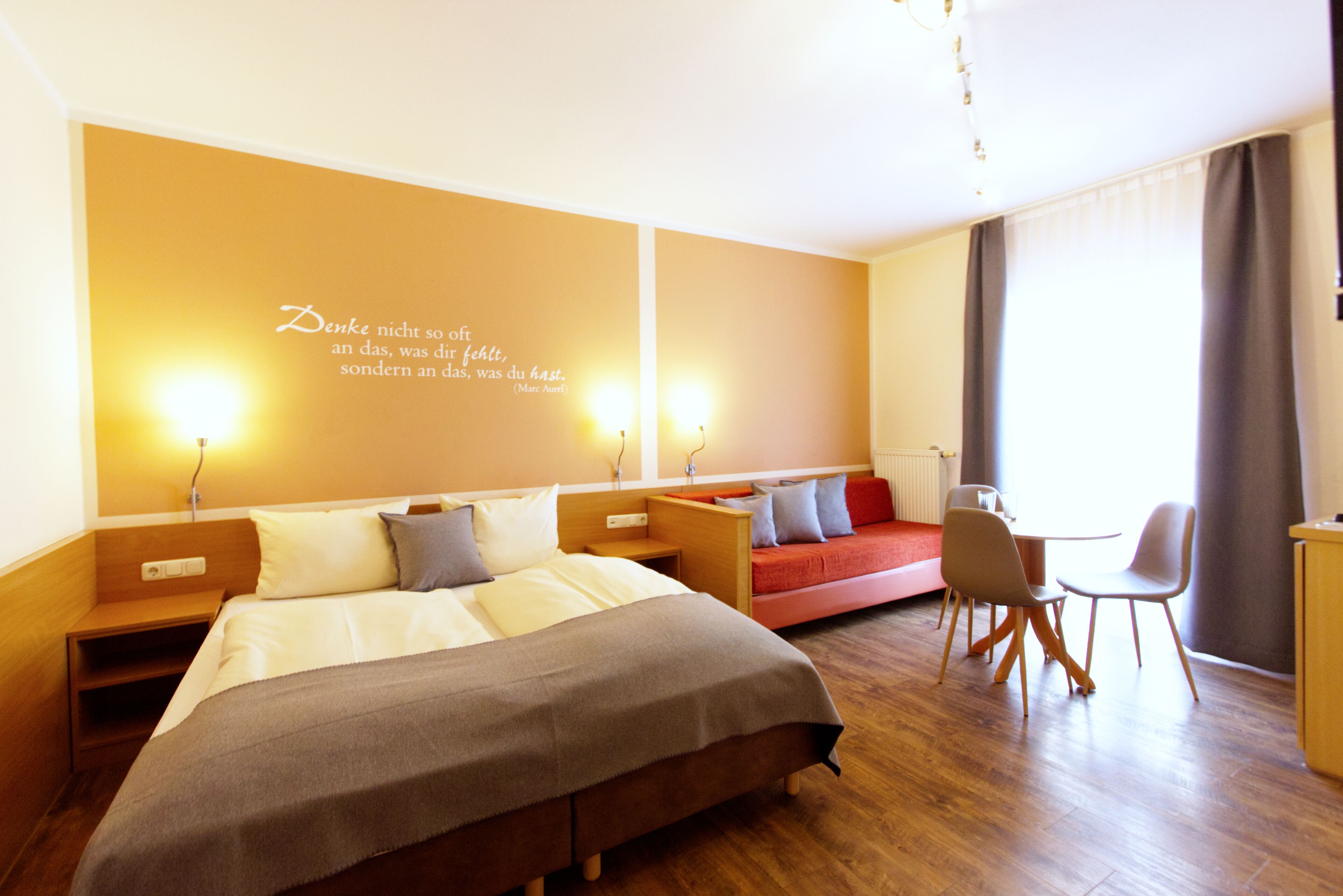 Een hotelkamer met een groot bed, een bank, een bureau en een stoel. Er is een muur met een citaat en twee wandlampen.