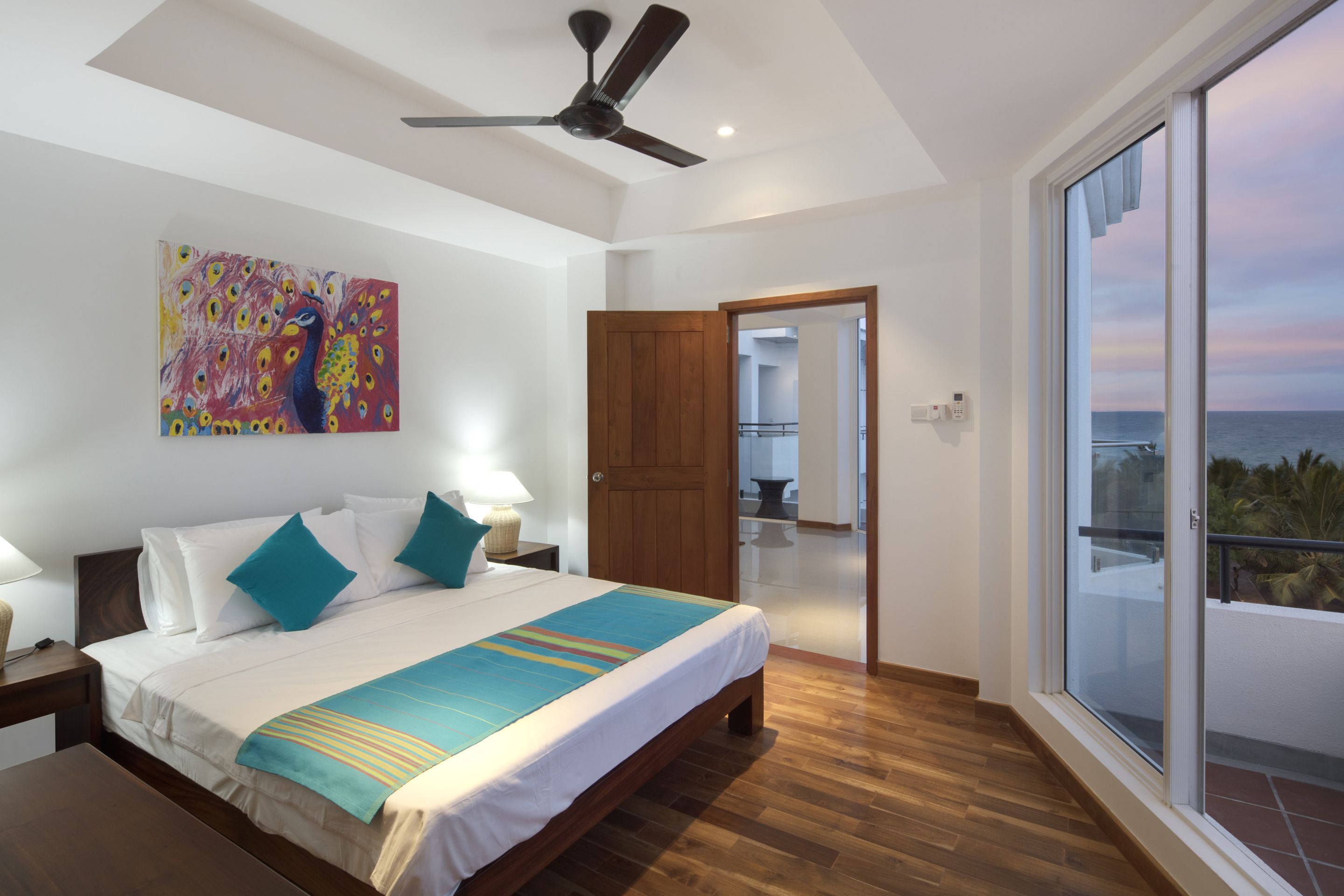 A bedroom with a bed, a ceiling fan, a painting, and a view of the sea through a balcony door.