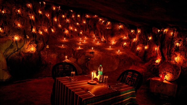 A cave dining area with tables set for two, candles, and a bottle of water. - Edakkal Hermitage (Sulthan Bathery)