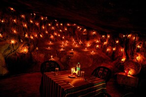 Un comedor en una cueva con mesas dispuestas para dos, velas y una botella de agua.
