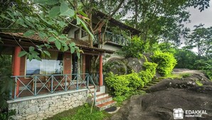 A house with a stone foundation, a balcony with blue railings, and lush greenery surrounding it. - Edakkal Hermitage (Sulthan Bathery)
