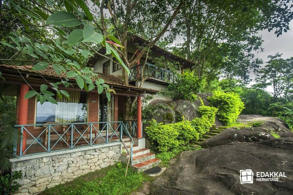 A house with a stone foundation, a balcony with blue railings, and lush greenery surrounding it.