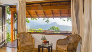 A room with two wicker chairs, a small table with fruit, and a large window offering a mountain view. - Edakkal Hermitage (Sulthan Bathery)