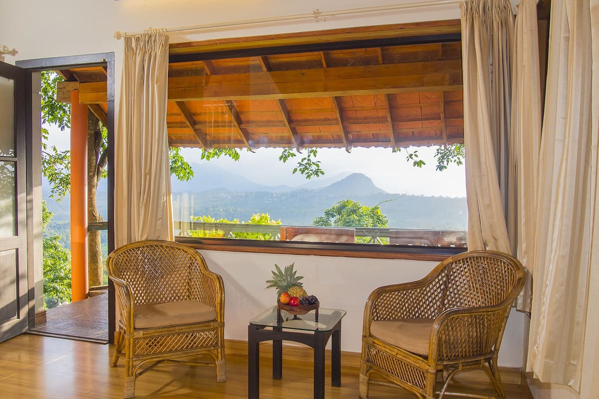 A room with two wicker chairs, a small table with fruit, and a large window offering a mountain view.